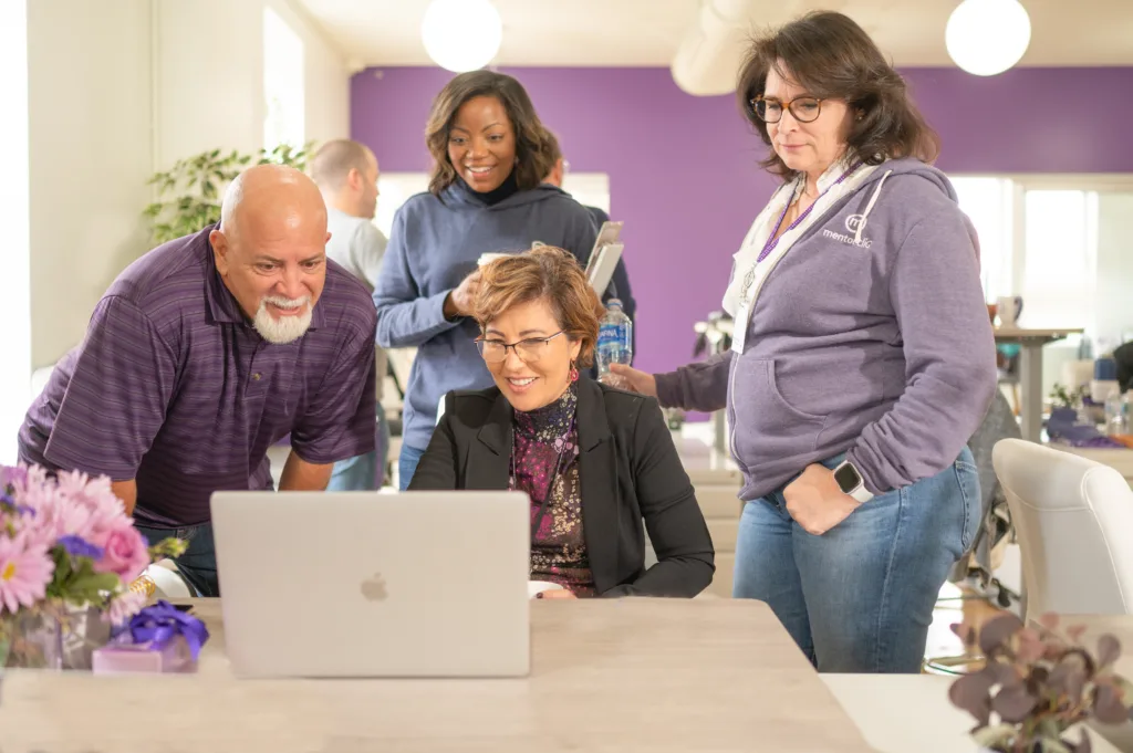 Feedforward vs Feedback: Is There a Difference? 3 Four colleagues collaboratively looking at a laptop in a brightly lit office decorated with purple accents and fresh flowers on the table.